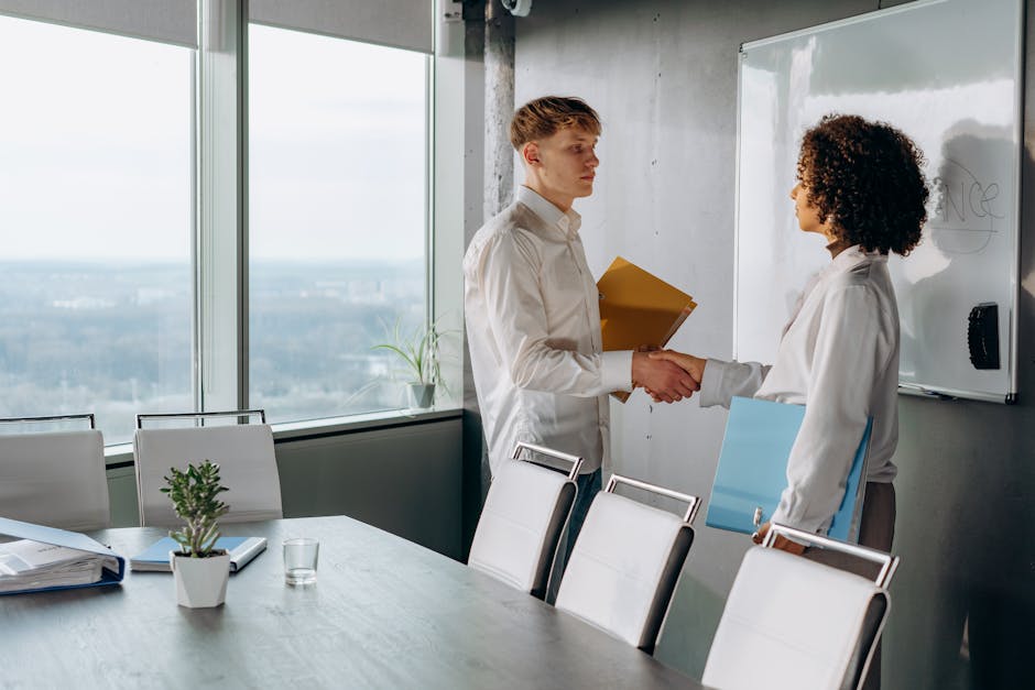 Two colleagues shaking hands in a bright conference room with documents.